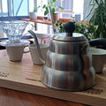 Stainless steel kettle on a wooden tray with coffee-making equipment in a cafe setting