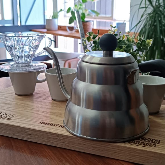 Stainless steel kettle on a wooden tray with coffee-making equipment in a cafe setting