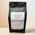 A 500g bag of Ambeáns Coffee Espresso Blend whole beans, in a matte black resealable pouch with a white and gold label, set against a textured beige wall.